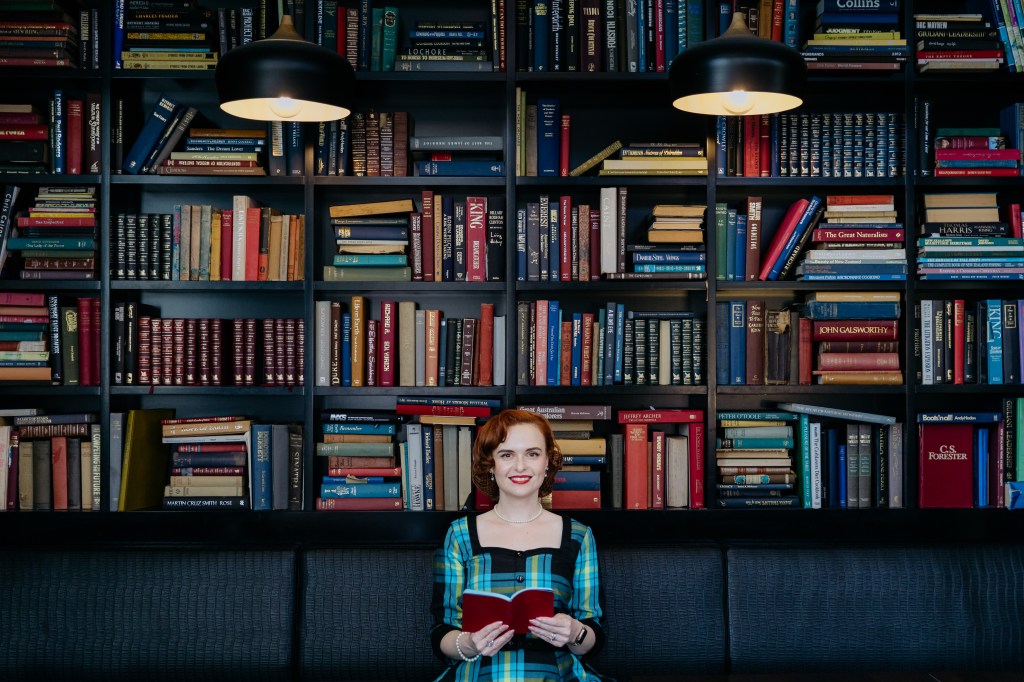 A woman holds open a book in front of a bookshelf